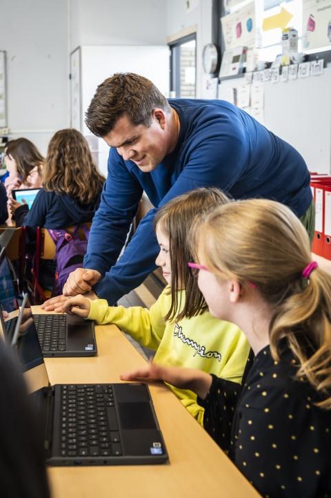 Meester helpt twee meisjes op de laptop in een klas van De Mozaïek.